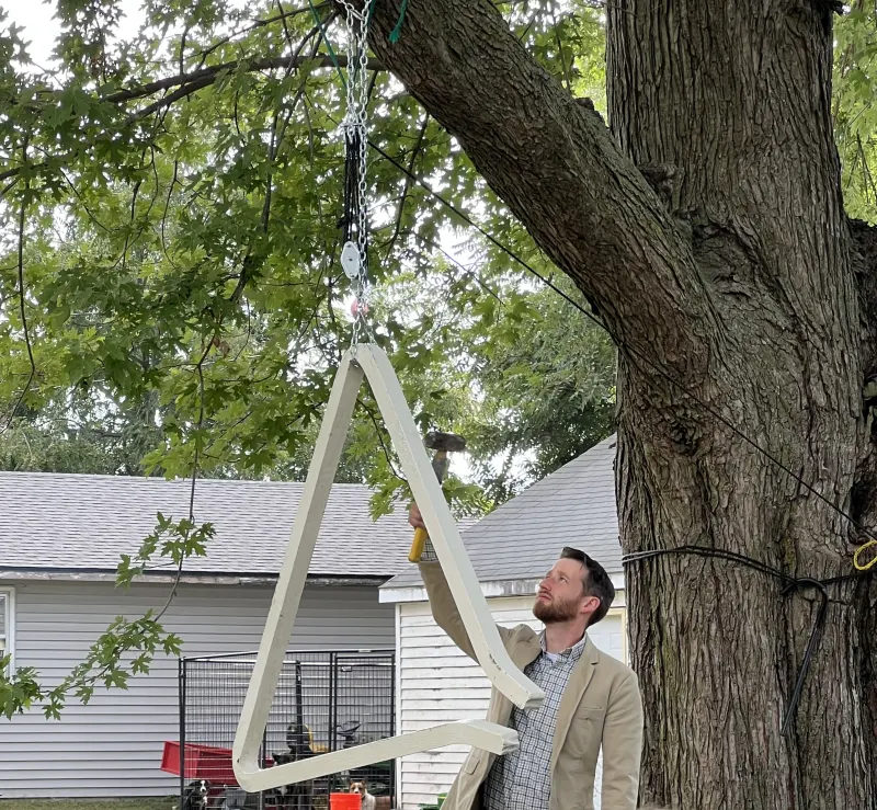 Pastor Jeremiah raises a hammer to ring the large white-painted iron triangle hanging by a chain from a tree branch